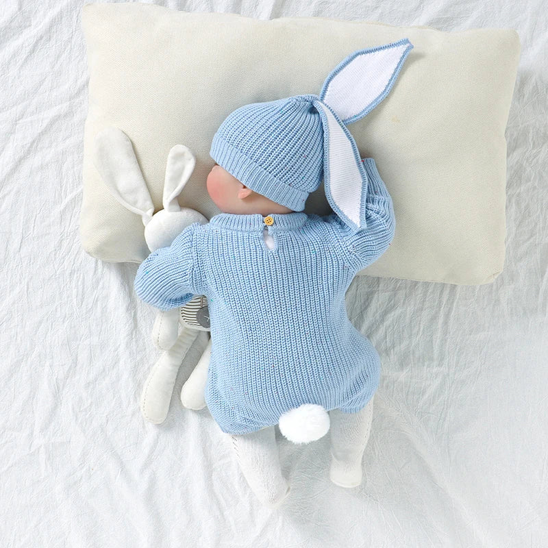 Baby in blue knitted bunny outfit lying on a white blanket with a plush toy.