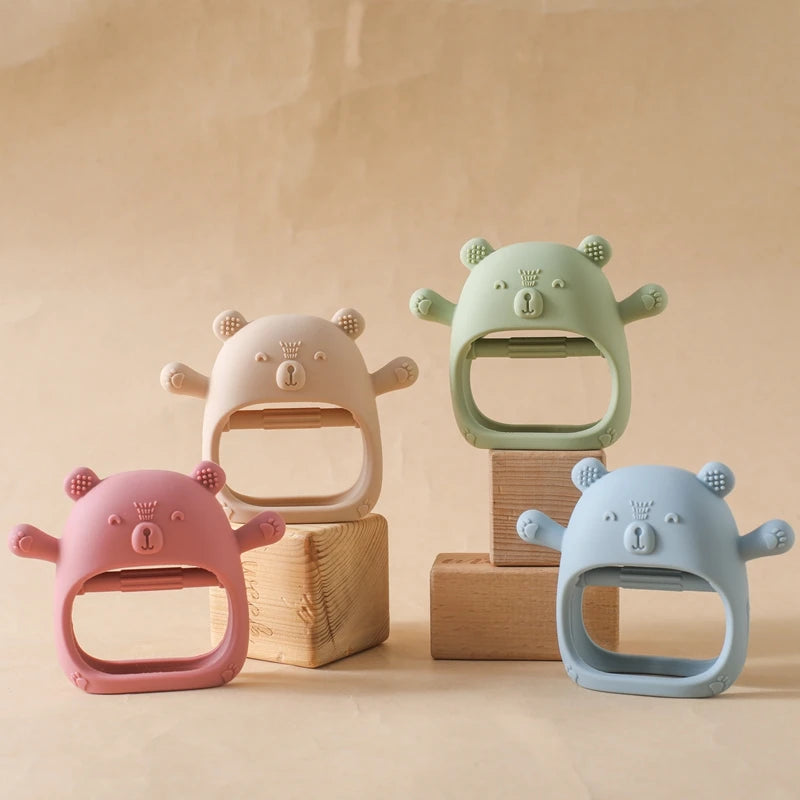 Four bear-shaped baby teething rings in pink, green, and blue on a beige background.