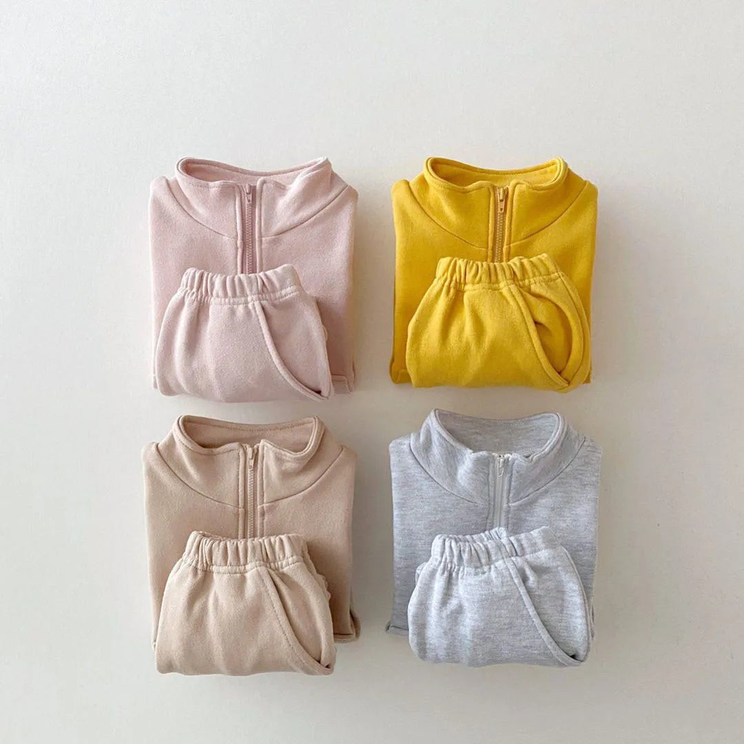 Four folded tracksuits outfits in pink, yellow, beige, and gray on a light background