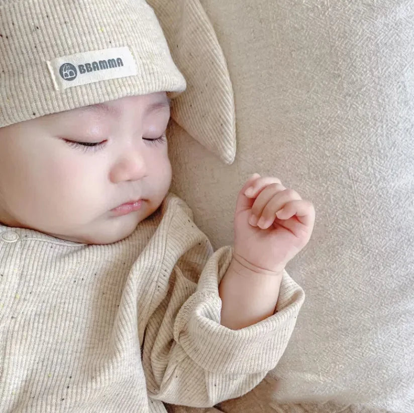 Baby wearing a beige hat with a brand label, lying on a soft surface.