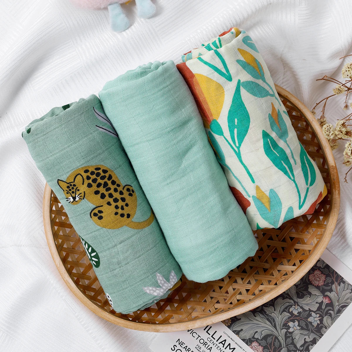 Three rolled-up fabric swaddles with animal prints in a woven basket on a white surface.