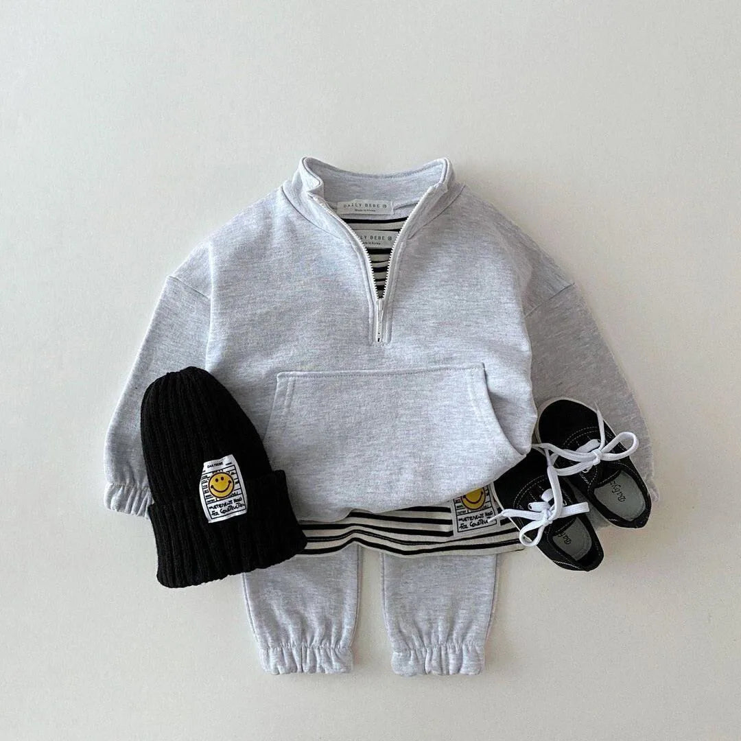 Children's outfit with gray half-zip sweatshirt, black beanie, striped socks, and black shoes on a light gray background.