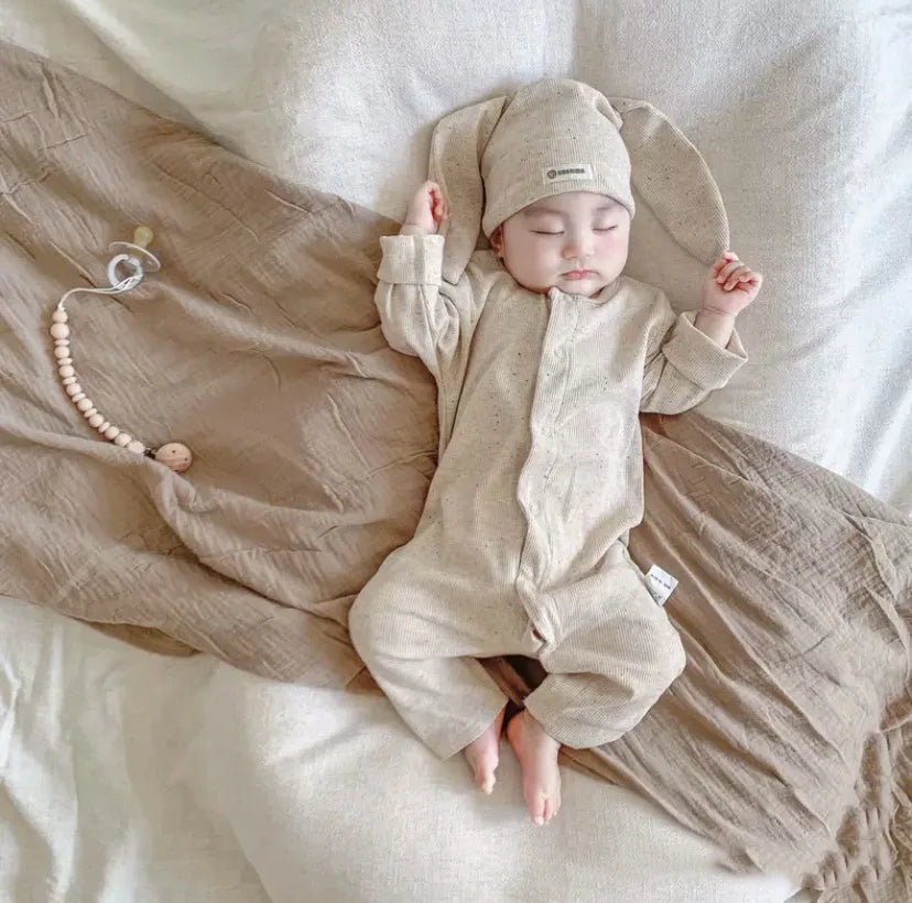 Baby in beige outfit and hat lying on a textured blanket with a teething toy nearby.