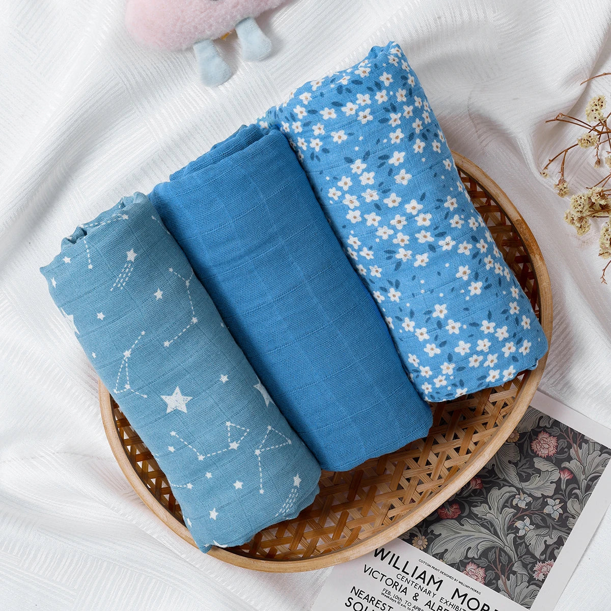 Three blue baby swaddle blankets with different patterns in a wooden basket on a white surface.