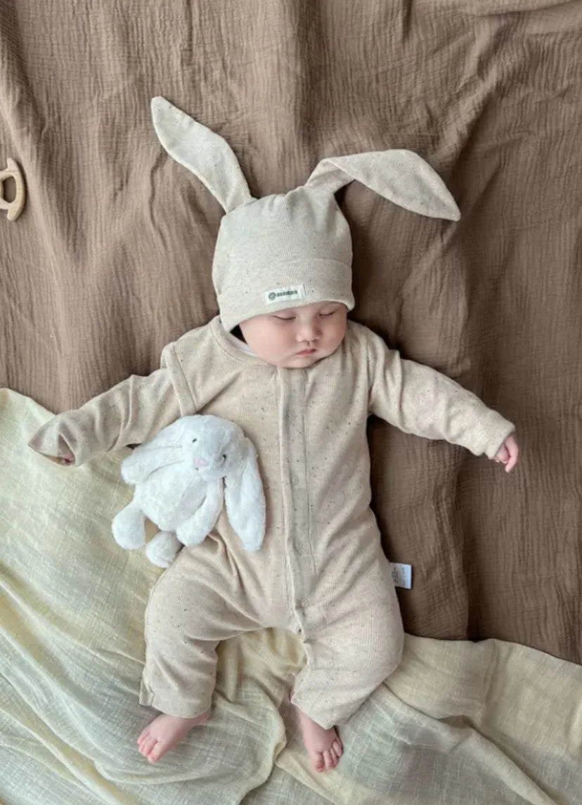 Baby in a beige bunny onesie with a white bunny plush toy on a brown blanket.