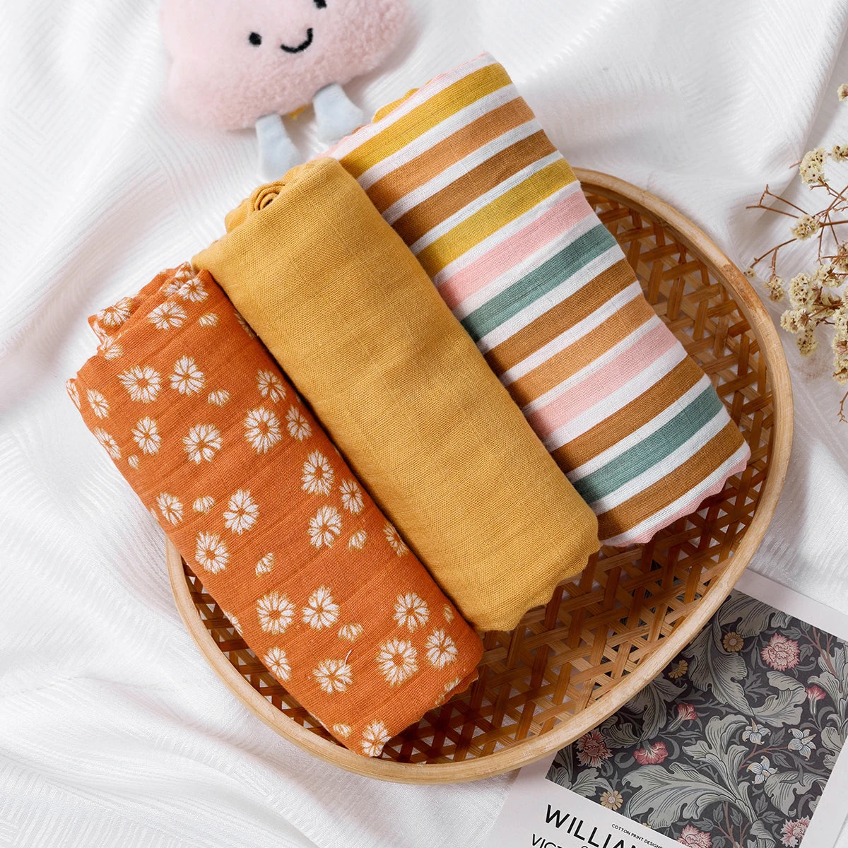 Three folded fabric swaddles in a wooden basket on a white surface with a small plush toy and dried flowers.