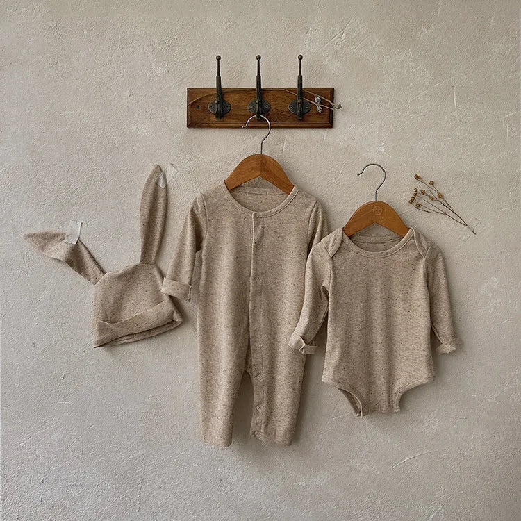 Beige baby romper and hat on hangers against a neutral wall.