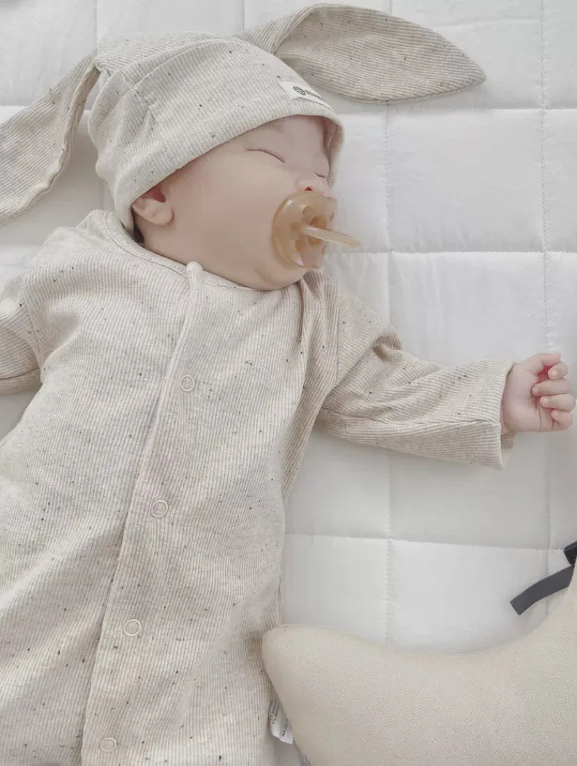 Baby in a beige outfit with bunny ears lying on a white quilted surface