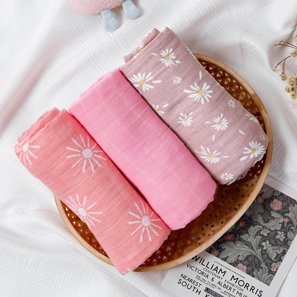 Three pink fabric swaddles with floral patterns in a wooden bowl on a white surface.